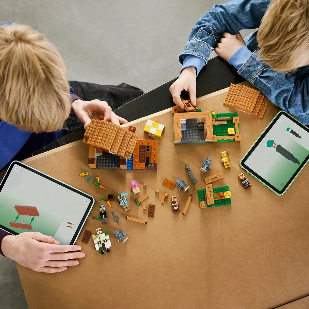 LEGO Minecraft Evoker Village Attack in brown, tan and green bricks is assembled by two children at a table, with loose pieces, figures and digital instructions nearby.