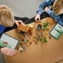 LEGO Minecraft Evoker Village Attack in brown, tan and green bricks is assembled by two children at a table, with loose pieces, figures and digital instructions nearby.