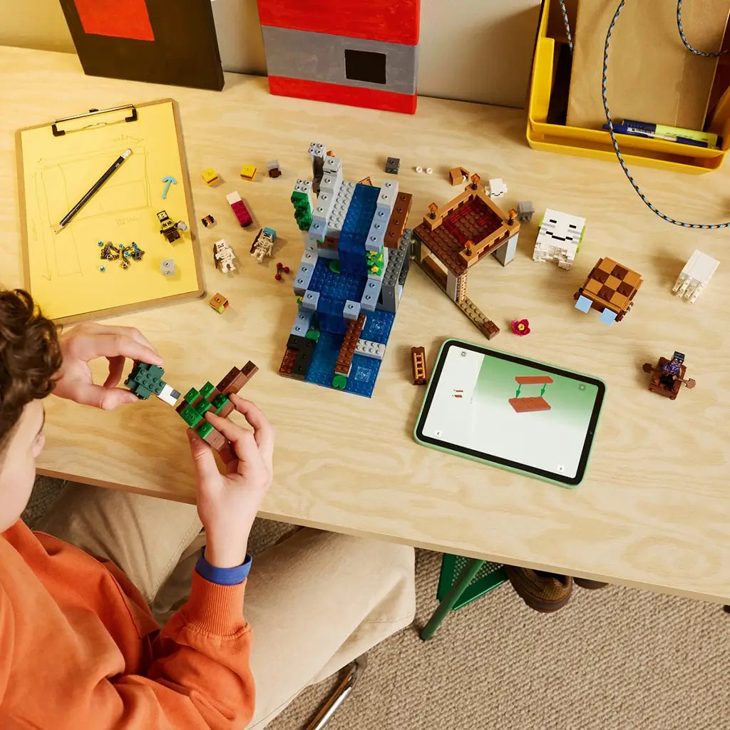 LEGO Minecraft Ghast Station in gray, brown, green and blue bricks is assembled by a child at a wood table, with loose pieces, figures and digital instructions nearby.