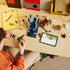 LEGO Minecraft Ghast Station in gray, brown, green and blue bricks is assembled by a child at a wood table, with loose pieces, figures and digital instructions nearby.