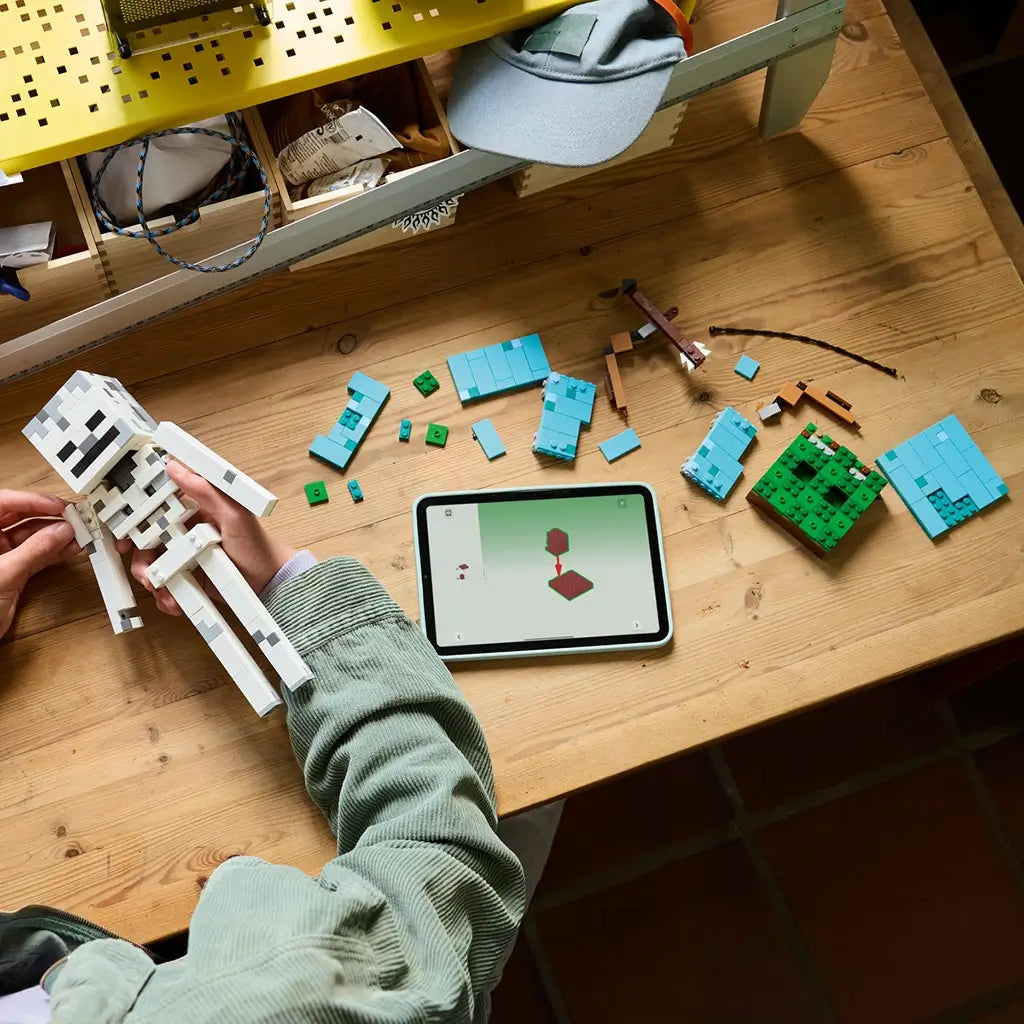 LEGO Minecraft The Skeleton build in white and gray bricks is held over a wooden table beside turquoise pieces, a brown bow and a tablet displaying digital building instructions.