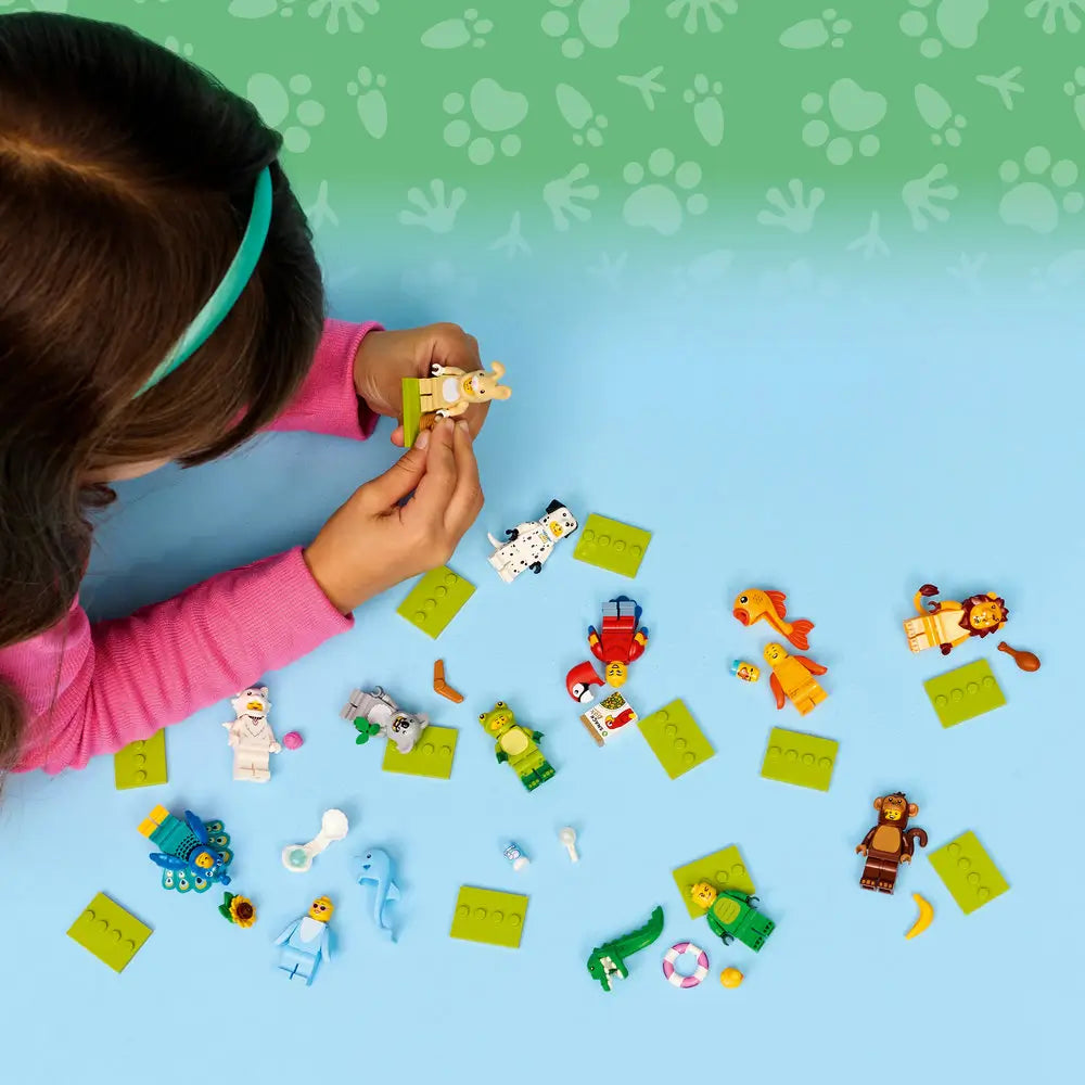 LEGO Minifigures Series 28 lifestyle image shows a child playing on a blue surface with scattered animal costume minifigures, green base plates, and accessories, viewed from above to highlight imaginative play.