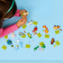 LEGO Minifigures Series 28 lifestyle build scene shows hands assembling and arranging animal costume minifigures on a blue tabletop, with loose parts, accessories, and green display plates.