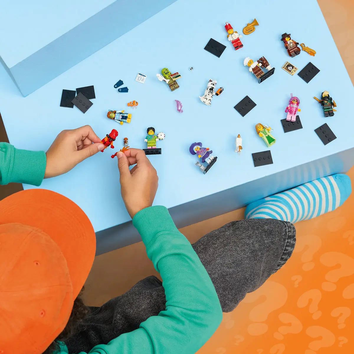 LEGO Minifigures Series 29 collectible toys rest on a pastel blue play surface while a child in an orange cap and green sweatshirt builds colorful plastic character figures.