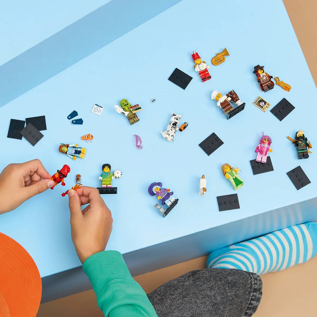 LEGO Minifigures Series 29 toys spread across a light blue tabletop with colorful plastic character figures, black display bases, and a child assembling pieces during play.