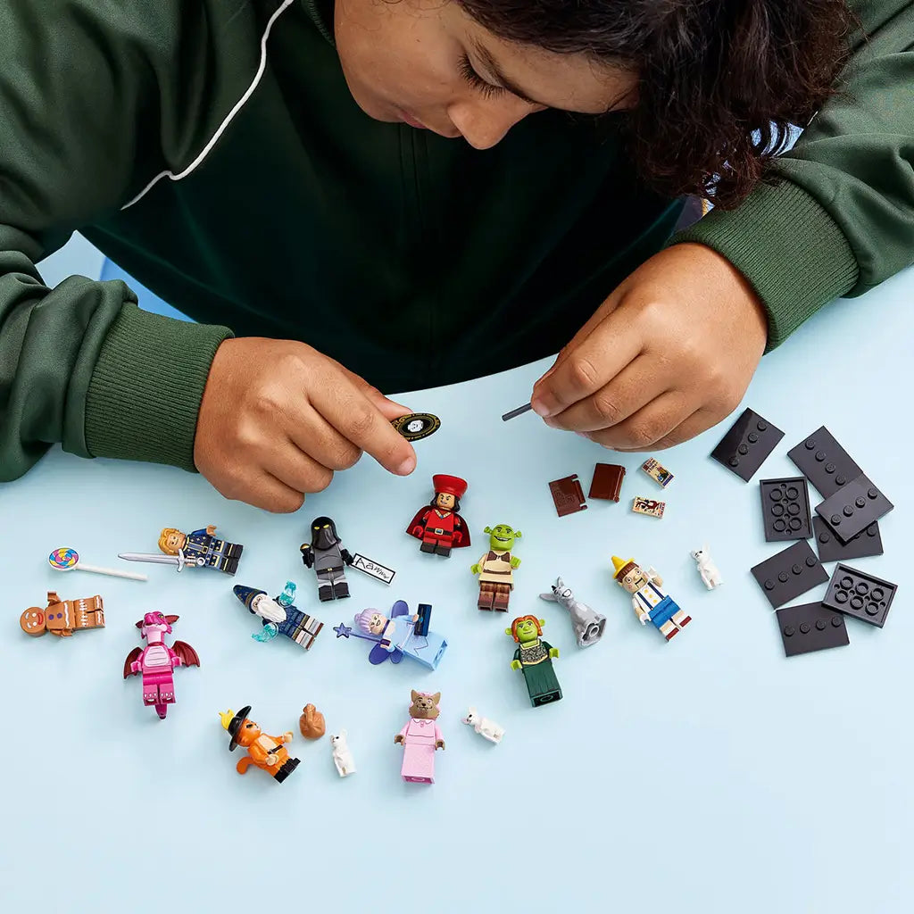 LEGO Shrek minifigures and accessories are spread across a light blue tabletop while a child assembles pieces, with black display bases grouped at the right.
