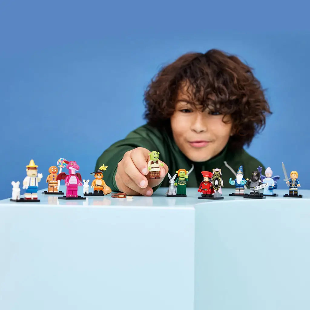 LEGO Shrek minifigures line a light blue display surface as a smiling child holds a Shrek figure toward the camera against a simple blue studio background.
