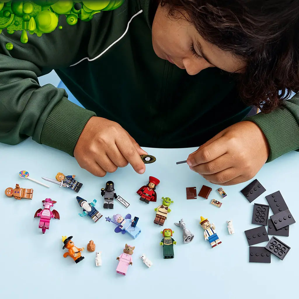LEGO Shrek minifigures are arranged across a light blue tabletop as a child in a dark green sweatshirt handles accessories and black display bases above the collection.