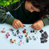 LEGO Shrek minifigures are arranged across a light blue tabletop as a child in a dark green sweatshirt handles accessories and black display bases above the collection.