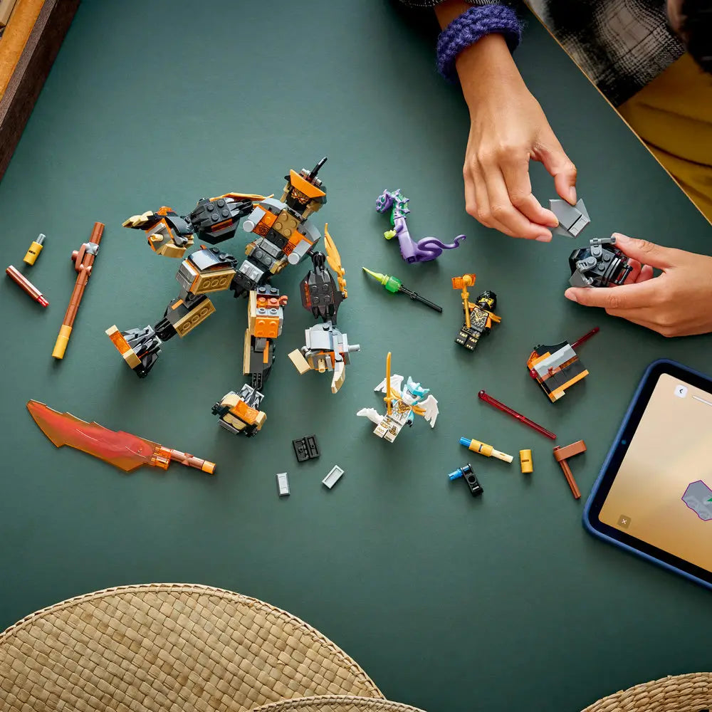 LEGO NINJAGO construction toy displayed during assembly on a green table, dark gray and gold mech body, scattered parts, ninja minifigures, and hands actively building for creative storytelling play.