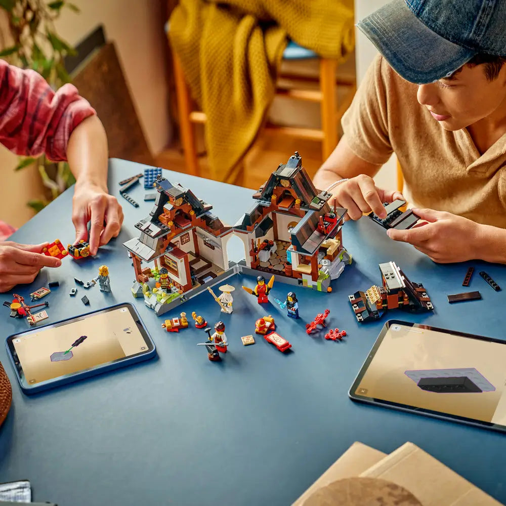 LEGO NINJAGO Four Weapons Blacksmith build shown mid-assembly in dark brown and gray plastic, with split building sections, loose bricks, minifigures, and tablet instructions arranged on a blue tabletop for guided group building play.