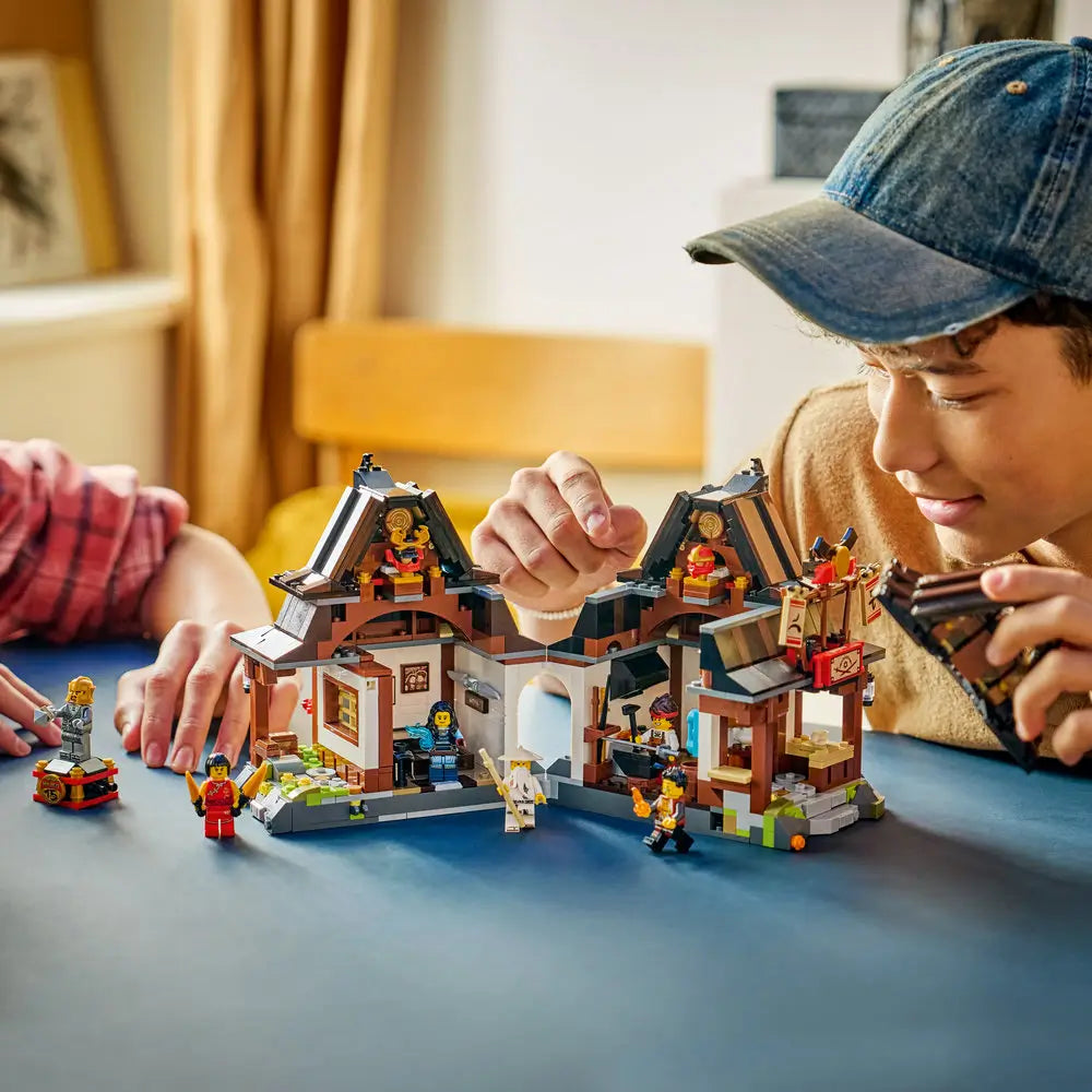 LEGO NINJAGO Legacy blacksmith set being assembled by two builders at a table, showing the modular building sections, scattered accessories, minifigures, and digital instructions to emphasize cooperative hands-on construction.