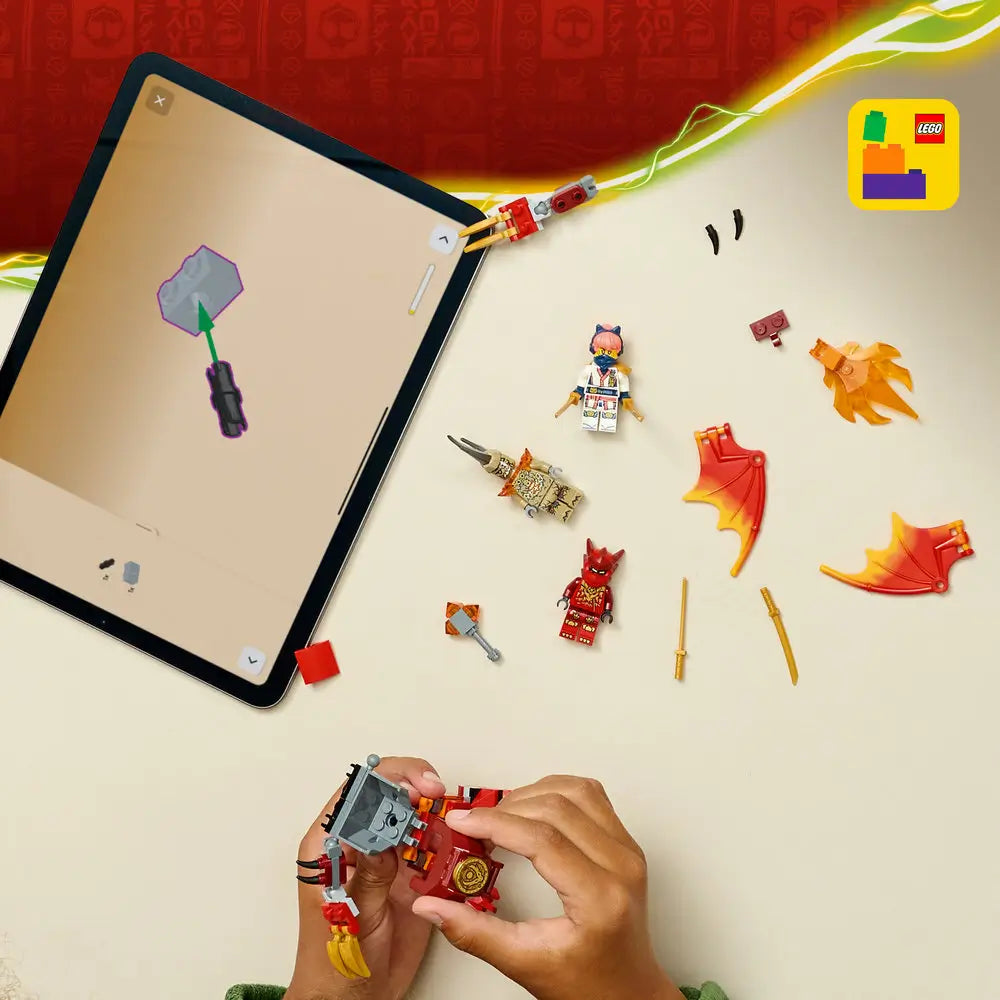 LEGO NINJAGO building set pieces in red, gold, and orange plastic are spread across a light tabletop as hands assemble a dragon mech, with a tablet showing digital instructions in the background.