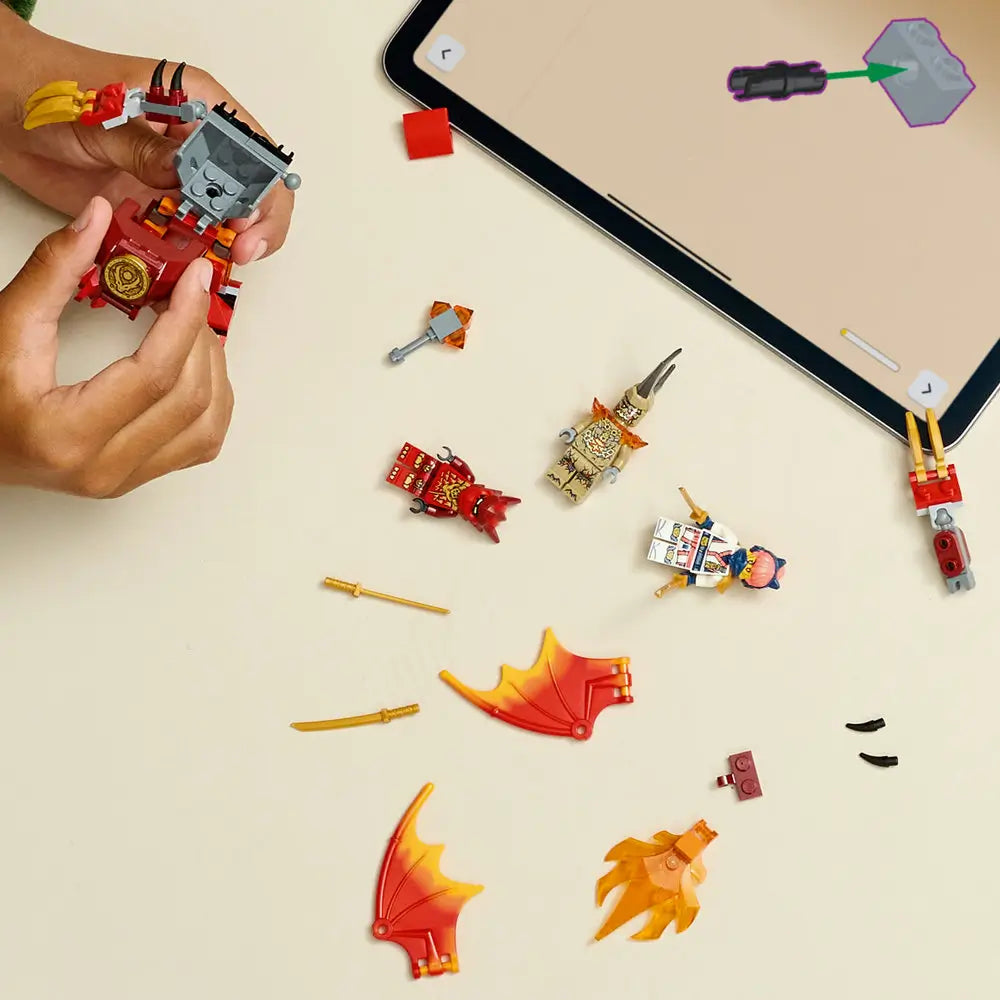 LEGO NINJAGO dragon mech parts in red, orange, and gold plastic lie on a neutral tabletop while hands build the model, surrounded by minifigures, weapons, wings, and a tablet with step by step instructions.
