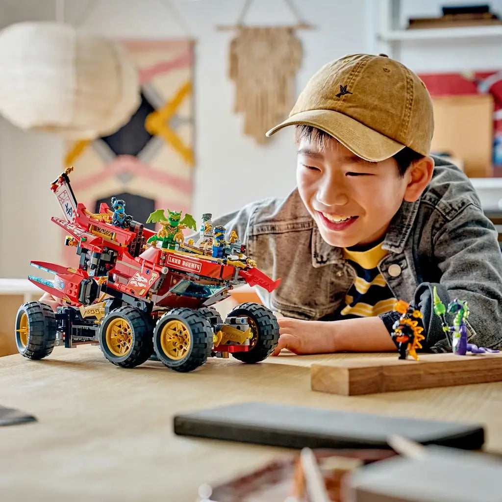 LEGO NINJAGO Land Bounty in red, black and gold plastic sits on six large wheels as a smiling builder plays with the vehicle and minifigures at a light wood table.
