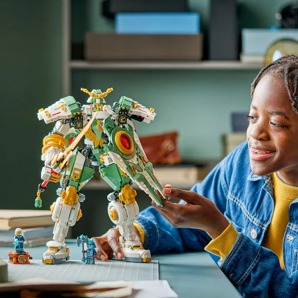 LEGO NINJAGO Titan Mech toy in green and gold plastic, large articulated robot form with sword and shield, action model being played with by a child at a desk in a bright indoor setting.
