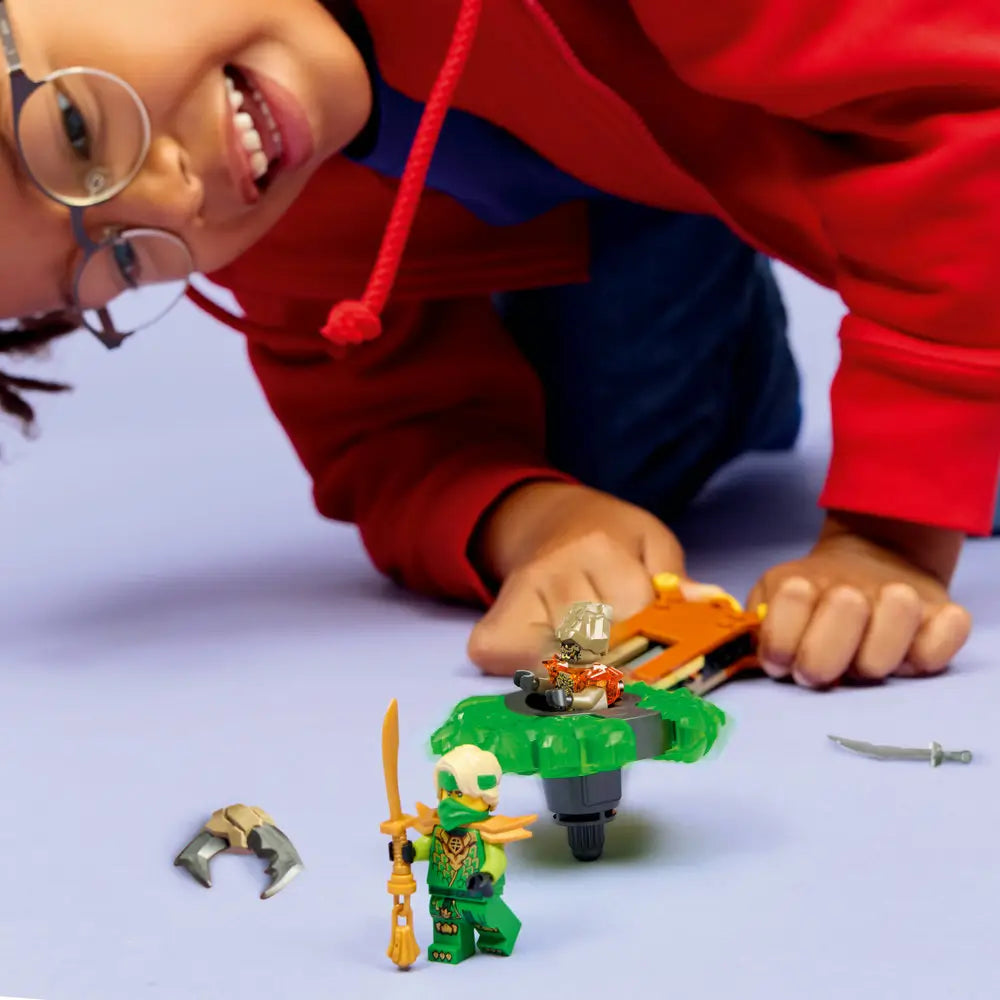 LEGO NINJAGO play scene shows a smiling child in a red hoodie leaning close while spinning a green plastic battle top with minifigures, emphasizing interactive play and imaginative storytelling.