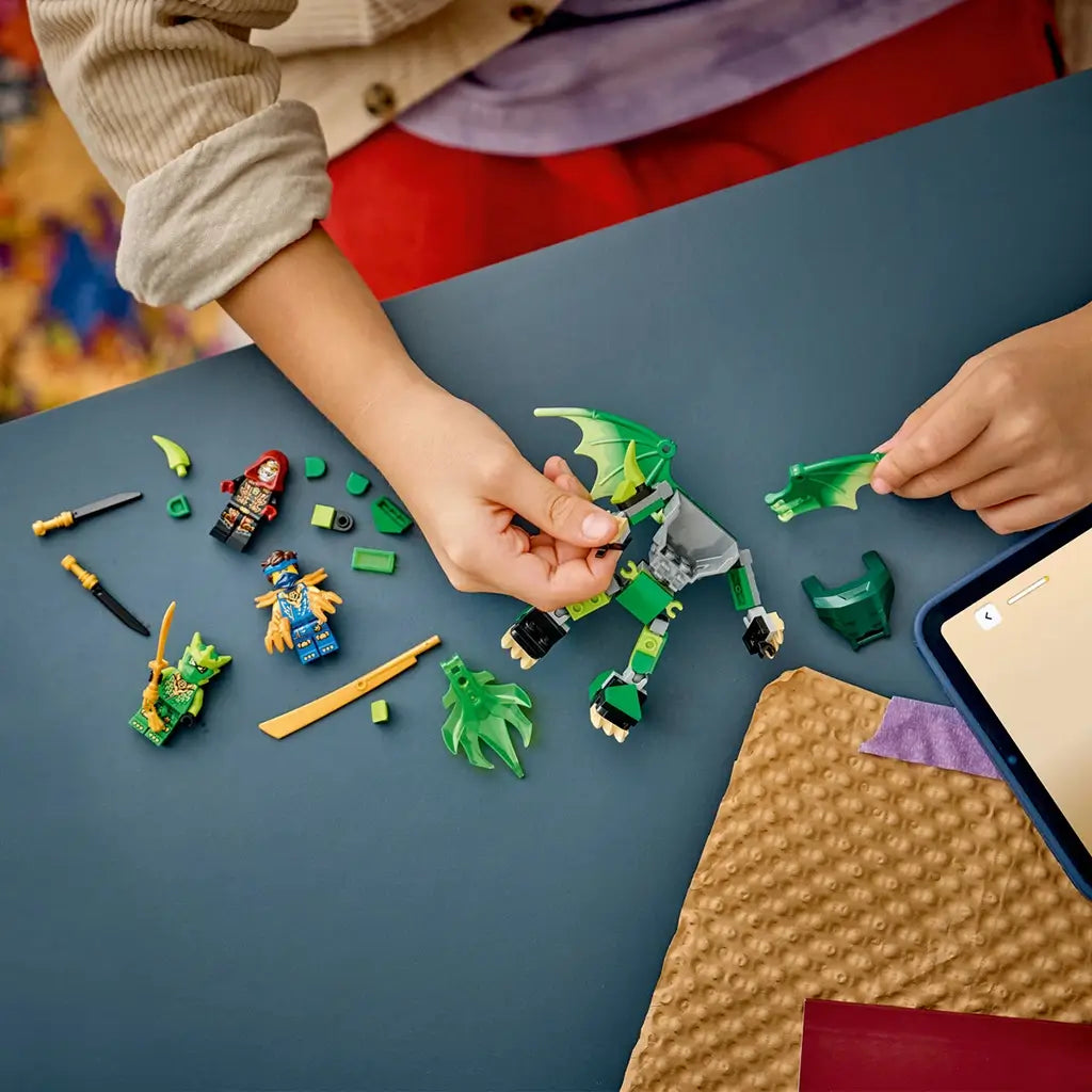 LEGO NINJAGO dragon mech set in green, gray and gold bricks is assembled by a child on a blue table, surrounded by loose pieces, weapons, and three minifigures.