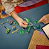 LEGO NINJAGO dragon mech set in green, gray and gold bricks is assembled by a child on a blue table, surrounded by loose pieces, weapons, and three minifigures.