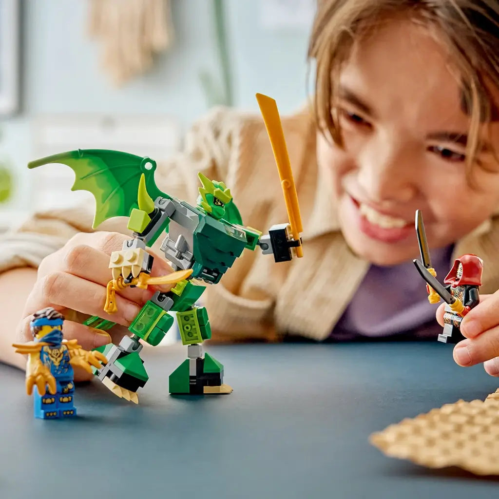 LEGO NINJAGO dragon mech toy in green, gray and gold bricks is held by a smiling child during play, facing a hooded villain while Lloyd stands on a blue table.