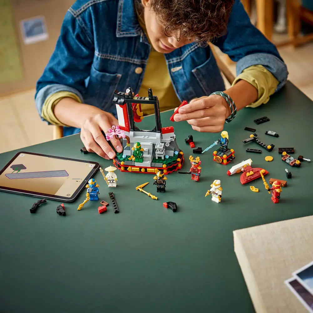 LEGO NINJAGO temple build in red, black, gray, and gold plastic shown mid-assembly, with loose bricks, ninja minifigures, and digital instructions arranged on a green tabletop for guided building play.