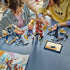 LEGO NINJAGO Twin Titan Mechs in red, blue, white and gold plastic are being assembled by two builders at a table with minifigures, loose parts and digital instructions.