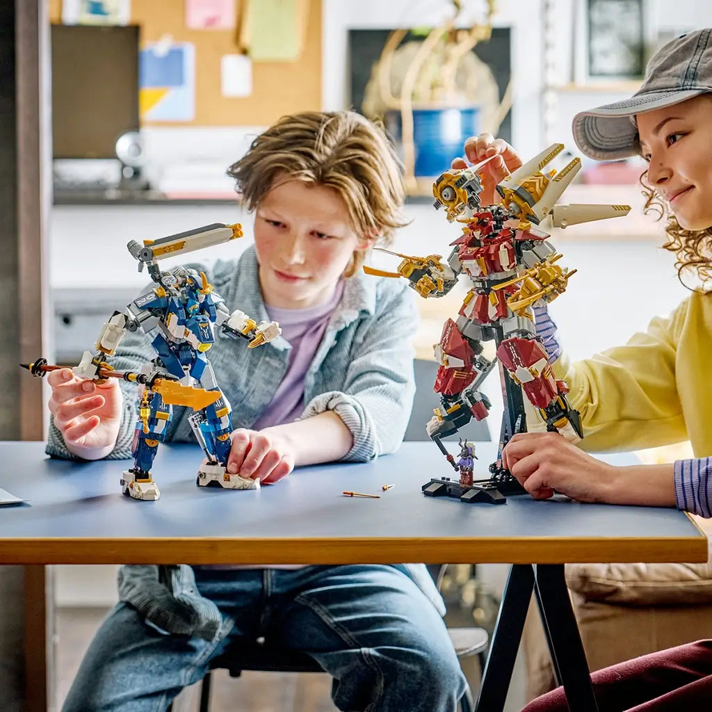 LEGO NINJAGO Twin Titan Mechs in red, blue, white and gold plastic are posed by two builders at a table, showing articulated limbs, gold weapons and display stands.