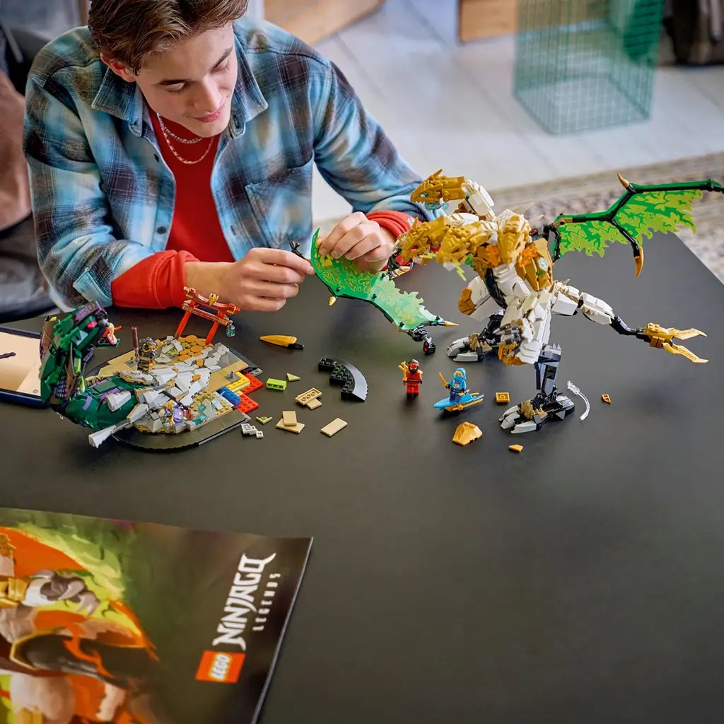 LEGO NINJAGO Ultra Dragon Battle in gold, white and green plastic is assembled by a builder at a dark table with the dragon, serpent, minifigures and loose bricks.