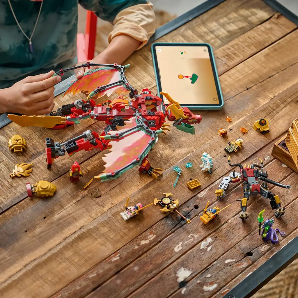 LEGO NINJAGO dragon set in red, gold and black plastic is arranged on a rustic wood table with minifigures and loose parts beside a tablet showing building instructions.