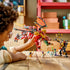 LEGO NINJAGO dragon mech in red and gold plastic is held aloft by a child, with flame wings extended above minifigures and a black-and-gold mech on a wooden table.