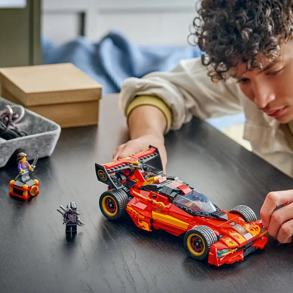 LEGO NINJAGO X-1 Ninja Charger in red, black and gold plastic is pushed across a dark tabletop by a builder, with sword-wielding minifigures positioned beside the car.