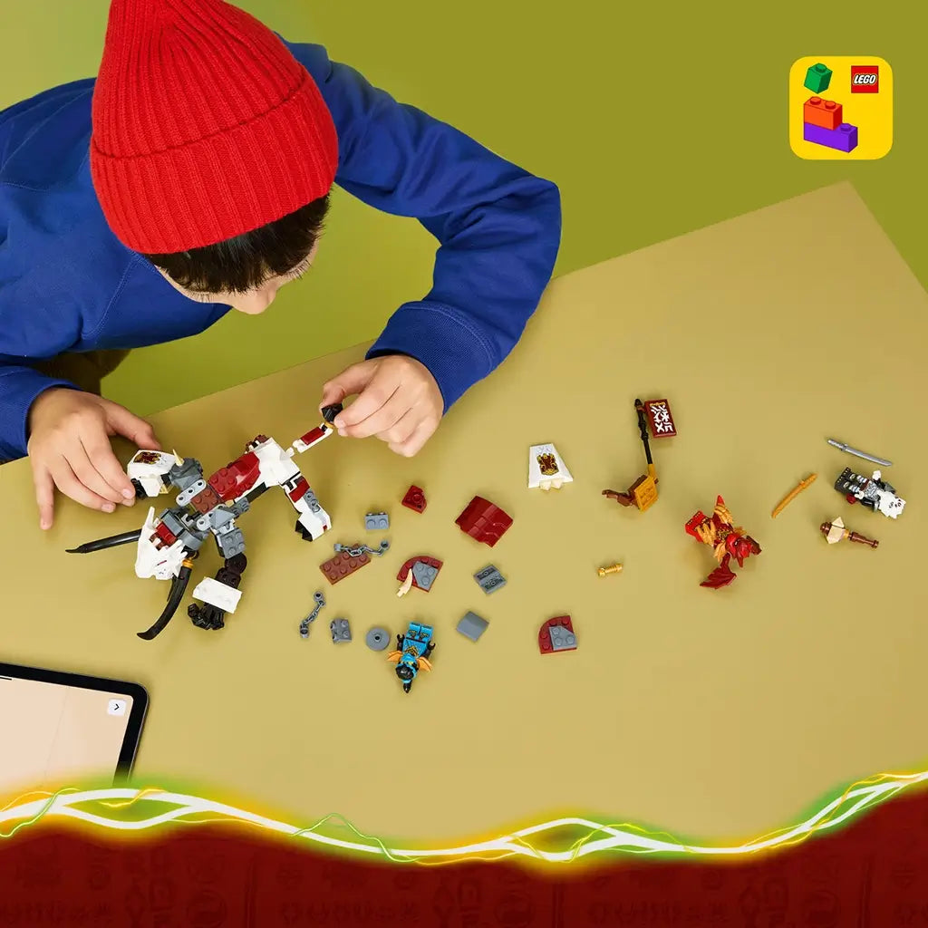 LEGO NINJAGO building set in white, black and red bricks is assembled on a tan table by a child in a red knit hat, with loose pieces, minifigures, and LEGO Builder icon.