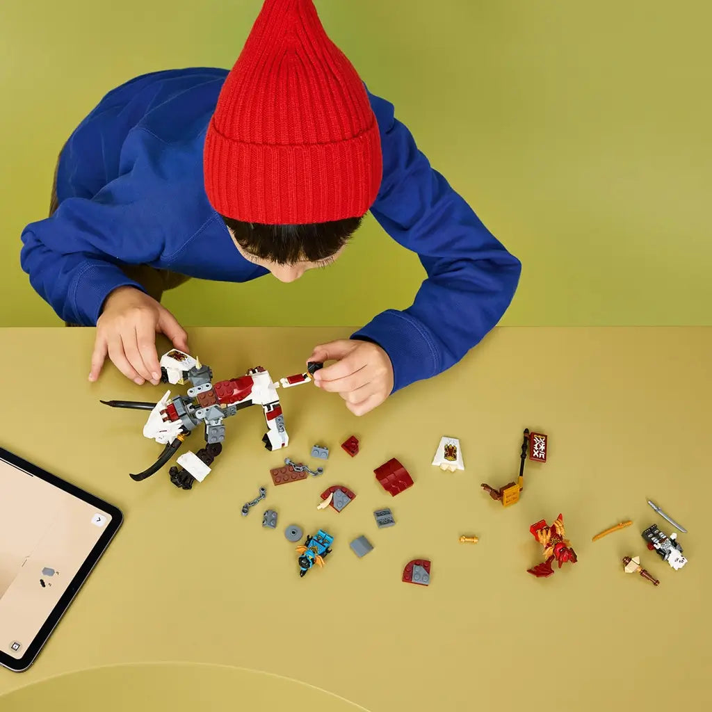 LEGO NINJAGO building set in white, black and red bricks is assembled by a child in a red knit hat and blue shirt, with loose pieces and minifigures on a tan table.