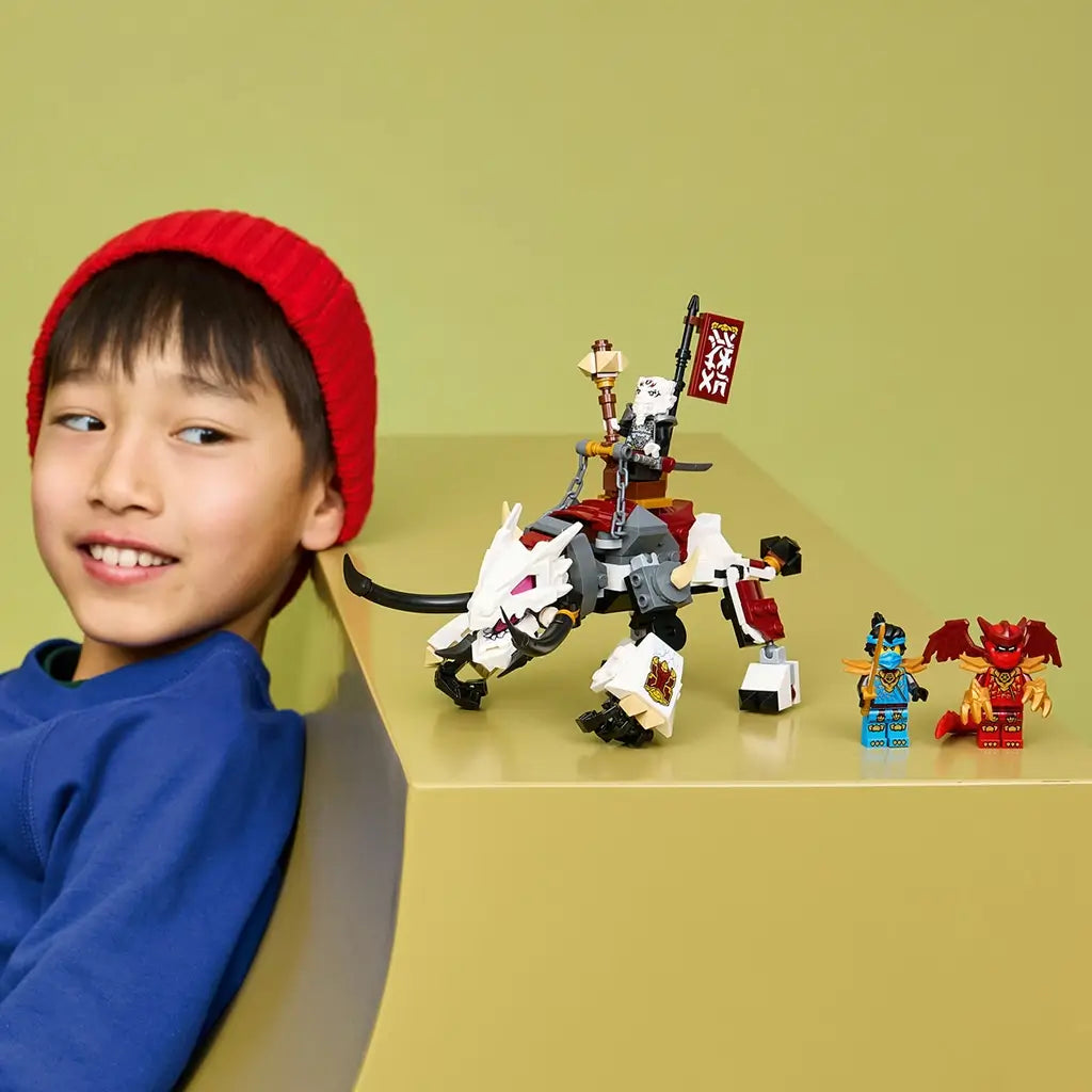 LEGO NINJAGO dragon toy in white, black and red bricks displayed with two minifigures on a tan table, beside a smiling child in a red knit hat and blue shirt.
