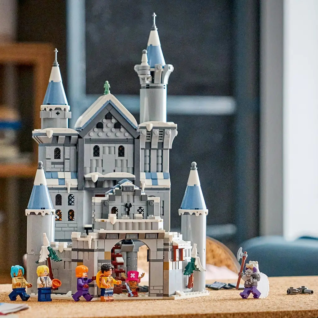 LEGO One Piece Battle at Drum Castle display features gray plastic walls, blue-gray turret roofs, snowy details, an arched gateway, and six minifigures on a cork tabletop.