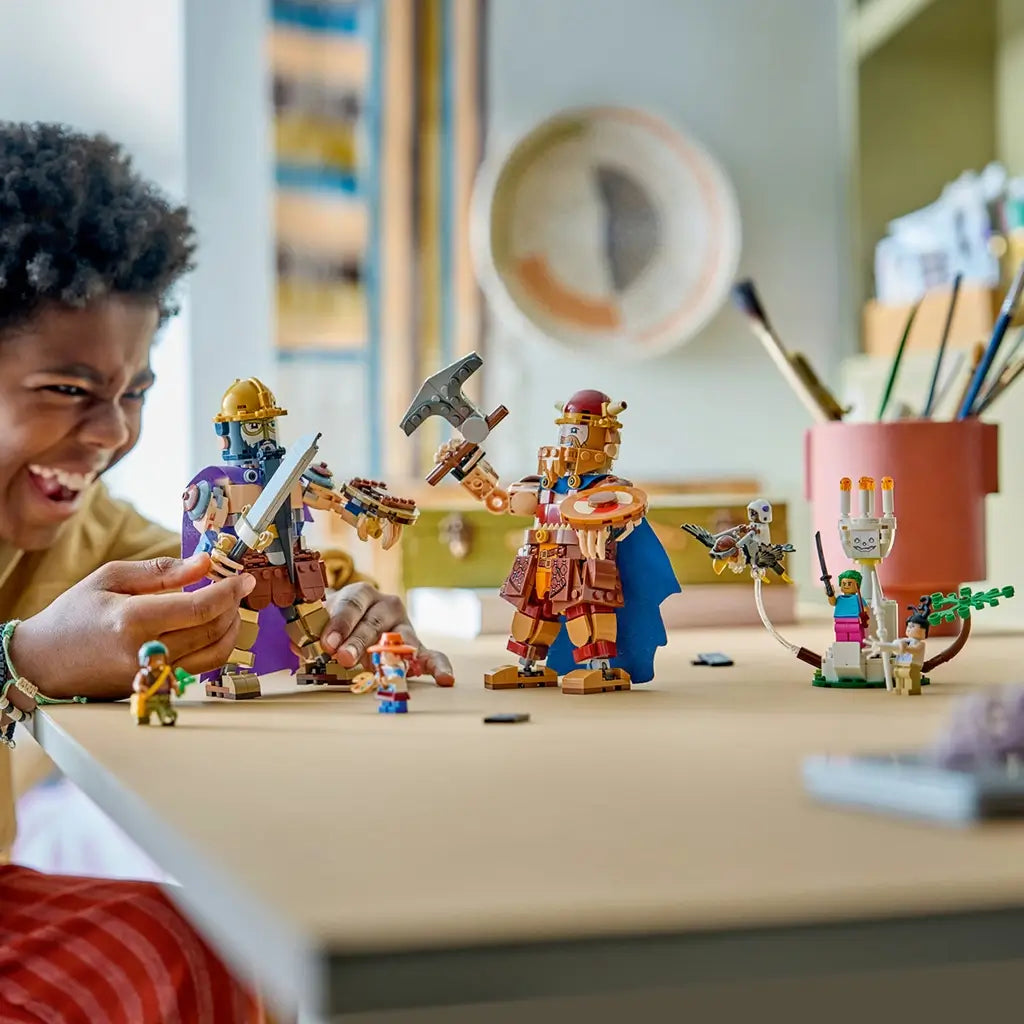 LEGO One Piece Dorry vs. Brogy play scene shows brown and tan brick-built giants facing off with weapons as a young builder moves figures across a tabletop indoors.