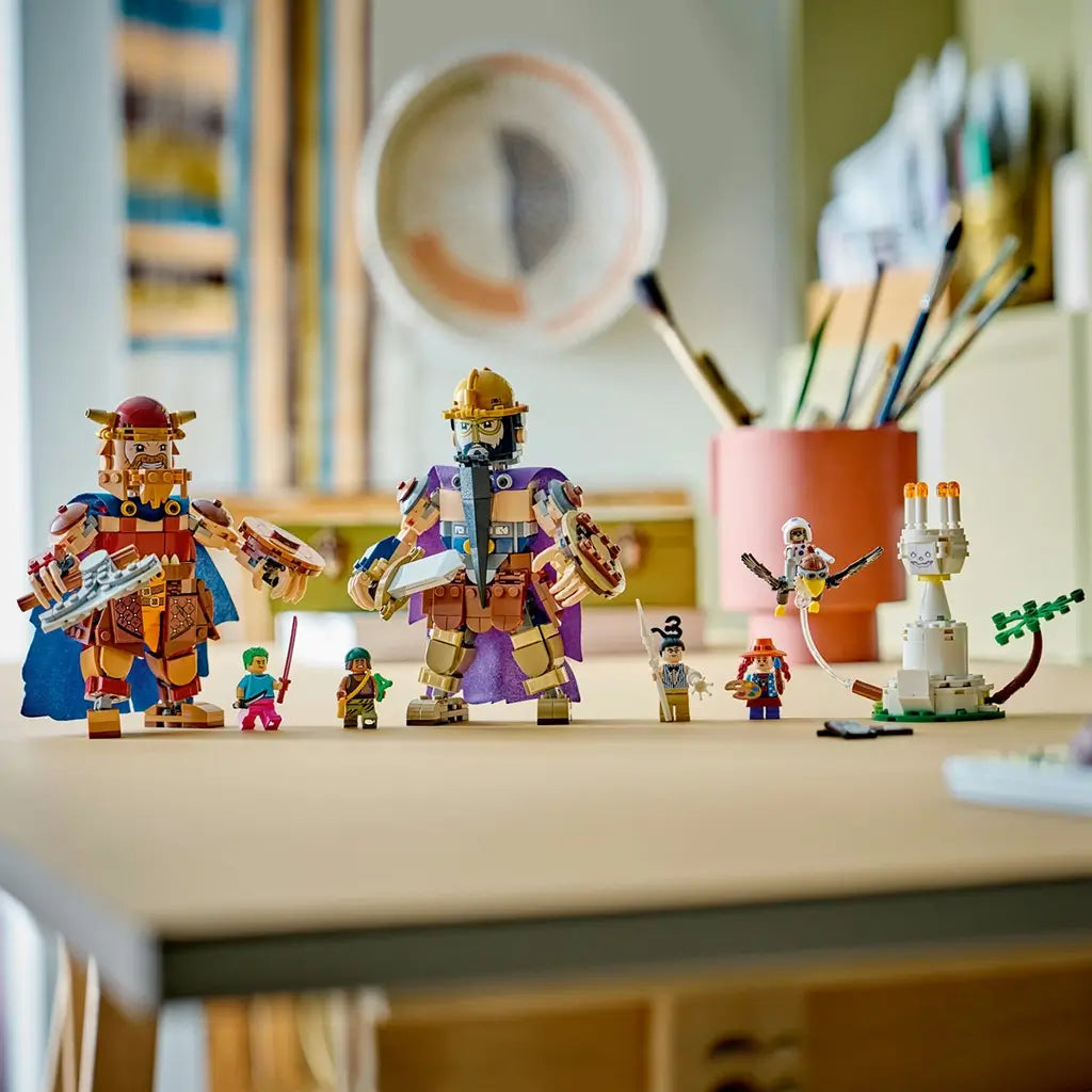 LEGO One Piece Dorry vs. Brogy display features brown and tan brick-built giants with capes, smaller figures, a white lookout structure, and palm tree on a tabletop.
