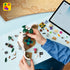 LEGO One Piece building set in white, brown and green plastic is assembled by a child at a blue table, with cave walls, flowering tree, loose pieces and tablet.