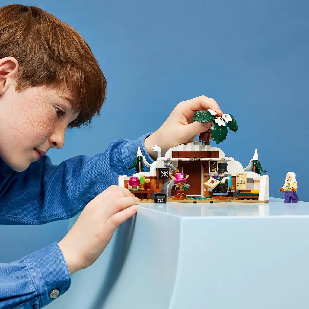 LEGO One Piece playset in white, brown and green plastic shows a child arranging the flowering tree above the snowy hideout, fireplace, bed and three minifigures.