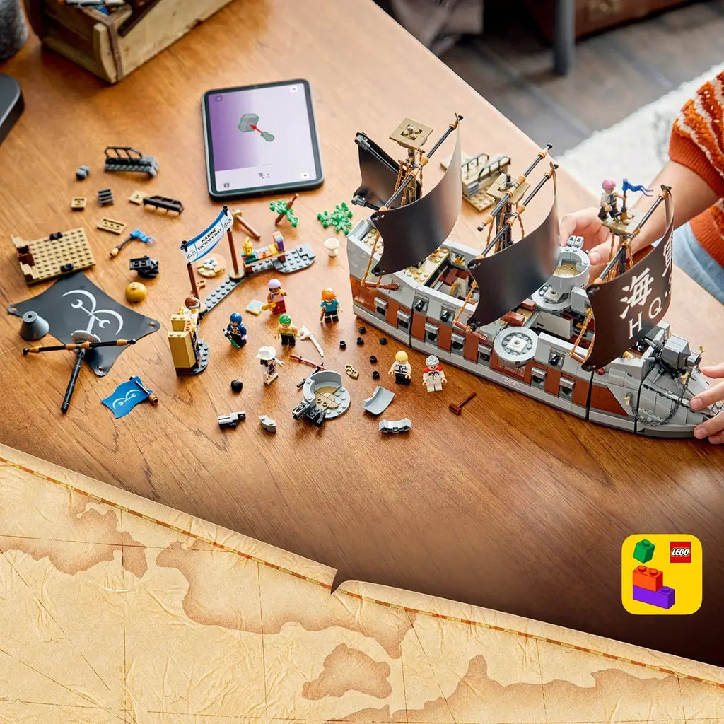LEGO One Piece Garp's Marine Battleship build in gray, brown, and black bricks sits partly assembled on a wooden table with loose pieces, minifigures, and a digital guide.