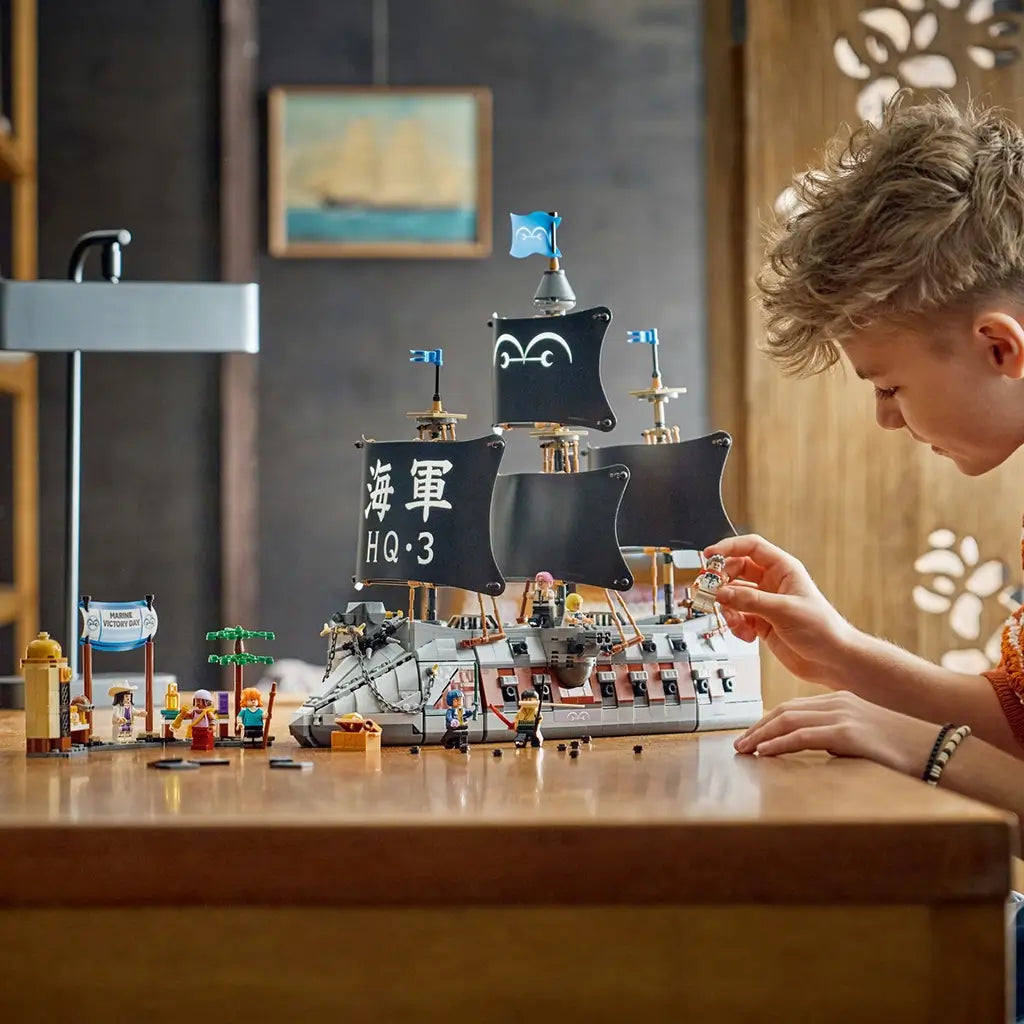 LEGO One Piece Garp's Marine Battleship in gray and brown plastic bricks has tall black sails and minifigures, while a young builder plays with the ship at a wooden table.
