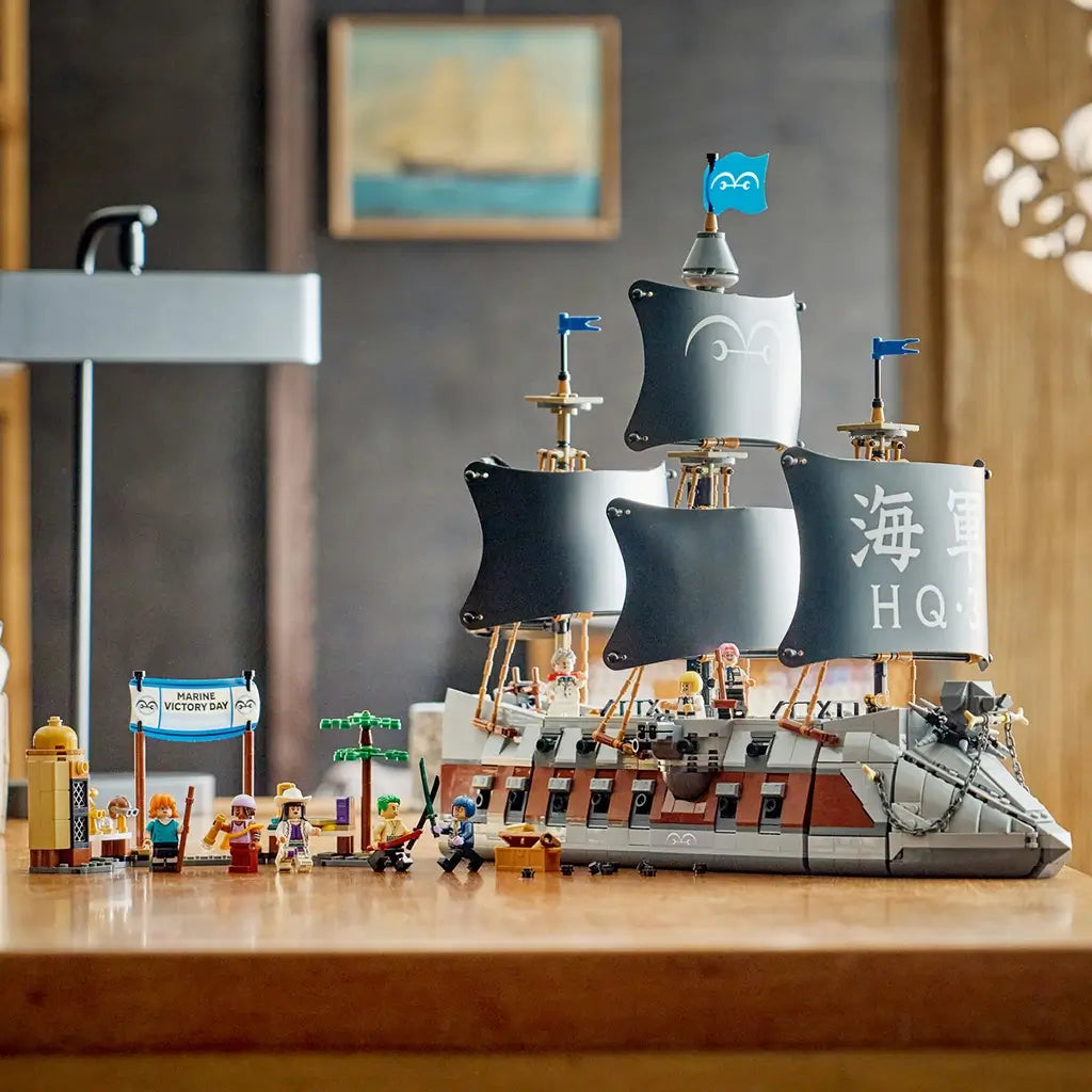 LEGO One Piece Garp's Marine Battleship display features a gray and brown brick ship with four black sails, blue flags, dock accessories, and minifigures on a wooden table.
