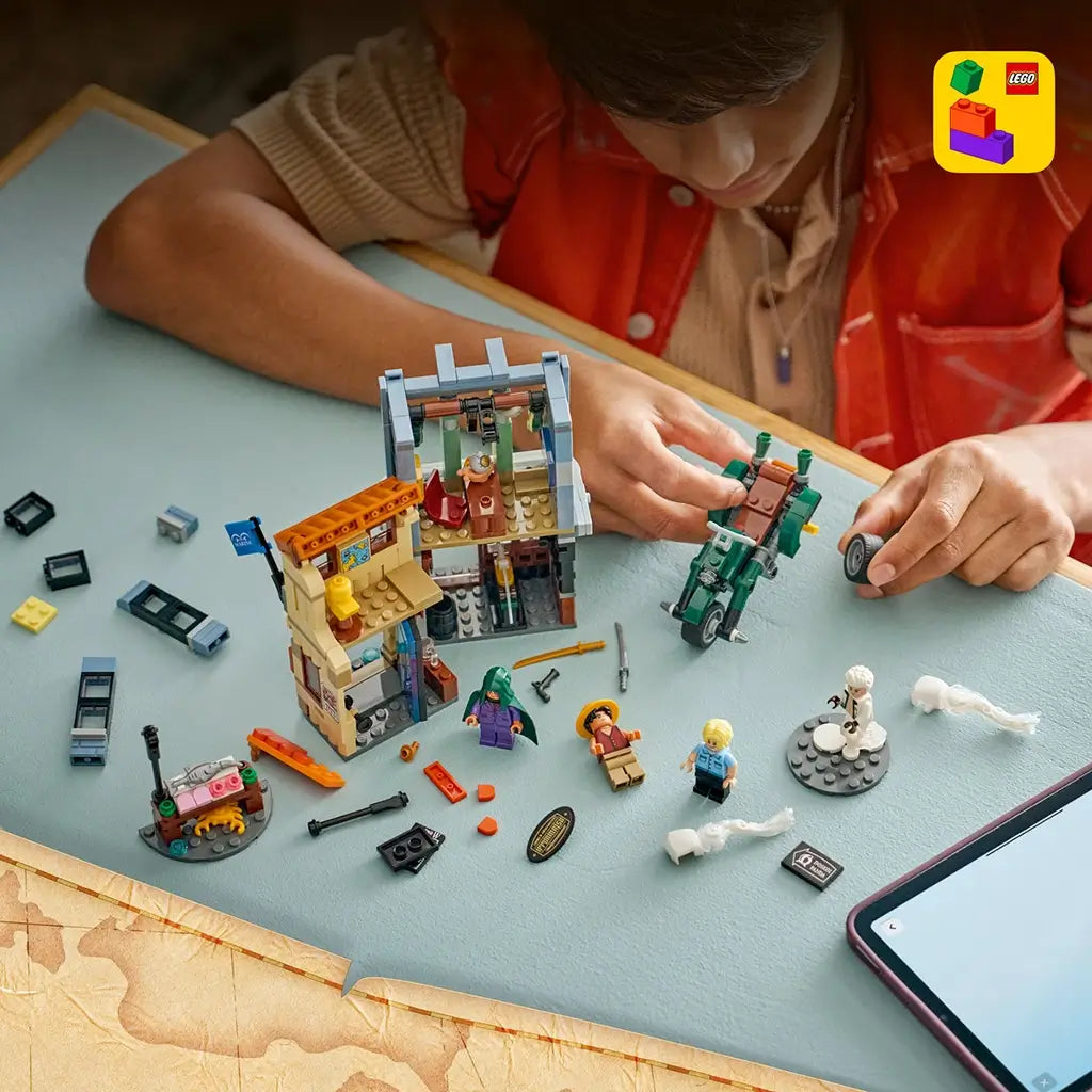 LEGO One Piece building set in multicolor plastic is assembled by a person at a blue table, with the Recruitment Center, motorcycle, minifigures and loose pieces.