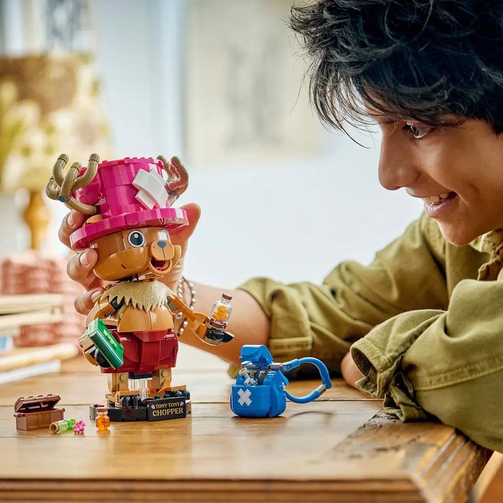 LEGO One Piece Tony Tony Chopper play scene shows a brown brick-built figure with pink hat, blue backpack, medicine accessories, and a smiling builder at a wooden table.