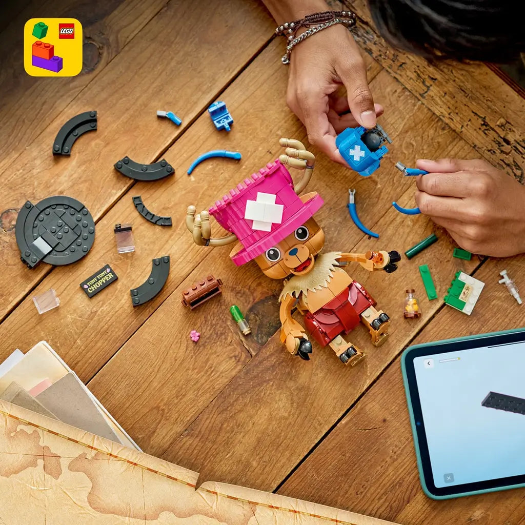 LEGO One Piece Tony Tony Chopper build scene shows a brown and pink brick-built figure, blue backpack parts, medicine accessories, loose pieces, and a tablet guide on wood.