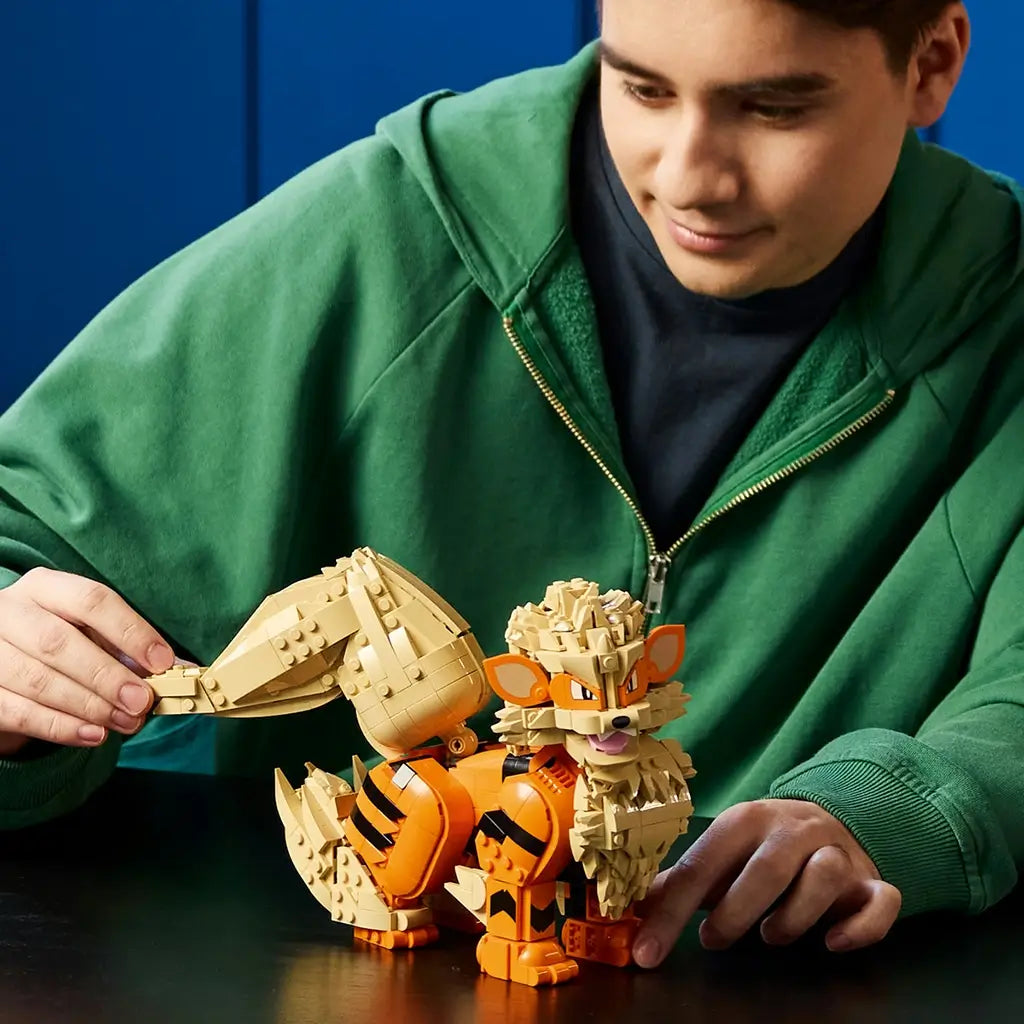 LEGO Pokémon Arcanine model in orange, tan, and black plastic bricks is adjusted by an adult builder, showing its large curled tail, furry mane, pointed ears, and paws.