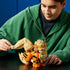 LEGO Pokémon Arcanine model in orange, tan, and black plastic bricks is adjusted by an adult builder, showing its large curled tail, furry mane, pointed ears, and paws.