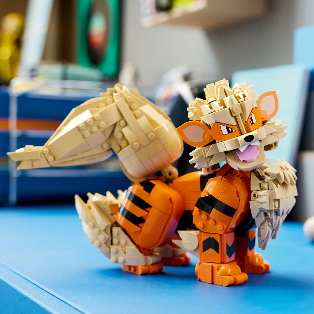 LEGO Pokémon Arcanine display in orange, tan, and black plastic bricks features a seated canine form with layered mane and curled tail, posed on a blue tabletop indoors.