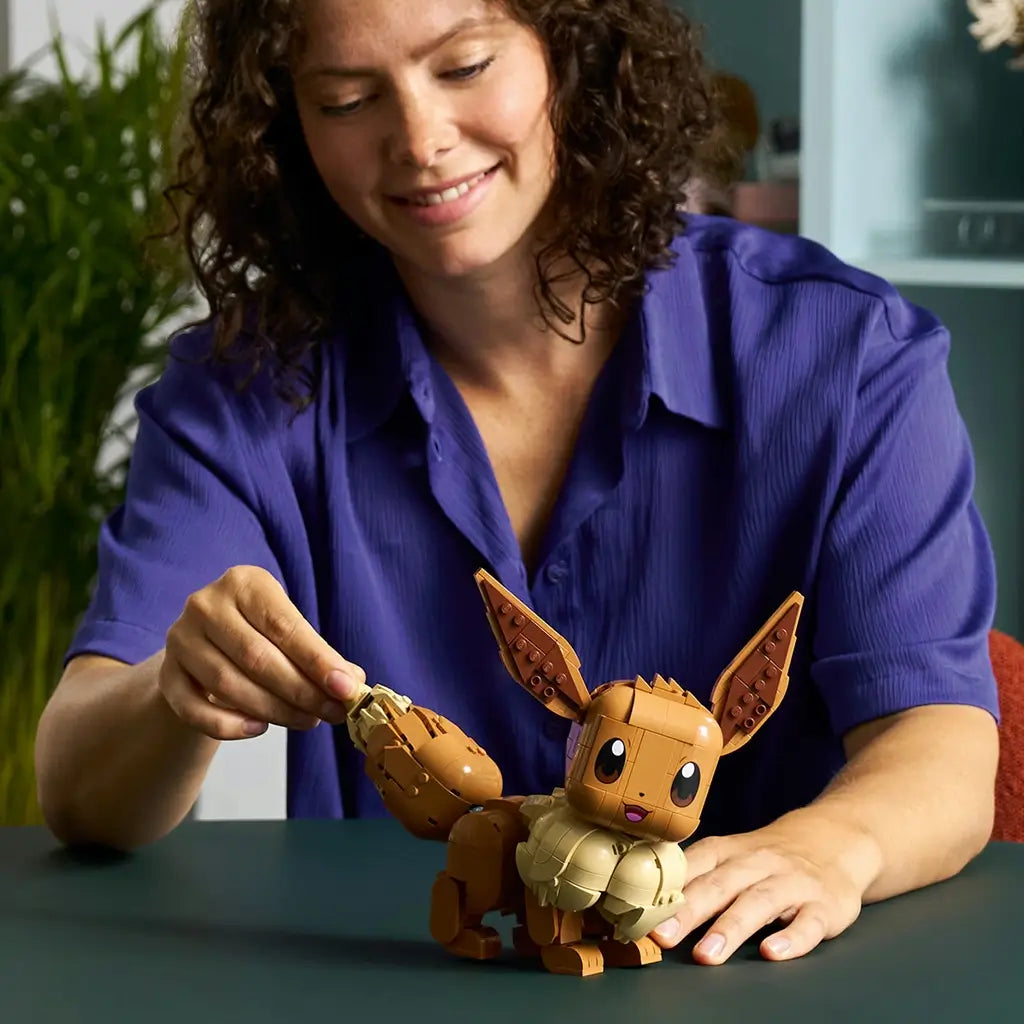 LEGO Pokémon Eevee model features brown and cream plastic construction, a compact animal form with articulated tail and ears, being adjusted by a builder at a tabletop in a home setting.
