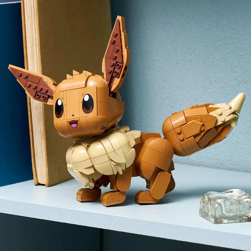 LEGO Pokémon Eevee display features warm brown and cream plastic surfaces, a rounded creature shape with large ears and curled tail, showcased on a shelf beside books and decorative glass accents.